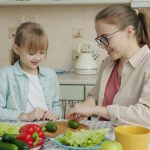 Young woman is cooking salad and feeding cute little daughter with cucumber slices in kitchen, family is talking and having good time together.